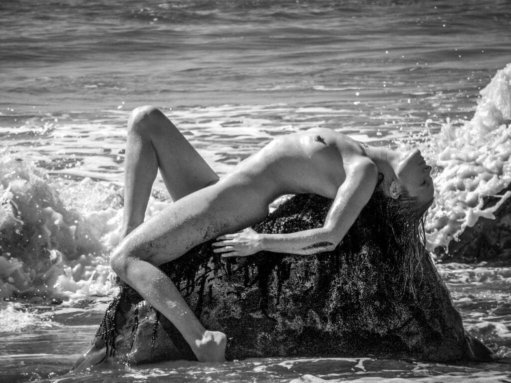 Black and white fine art nude reclining on rock as waves crash