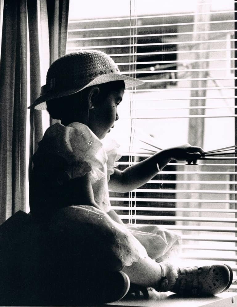 Child in a sun hat sitting by a window and looking through the blinds